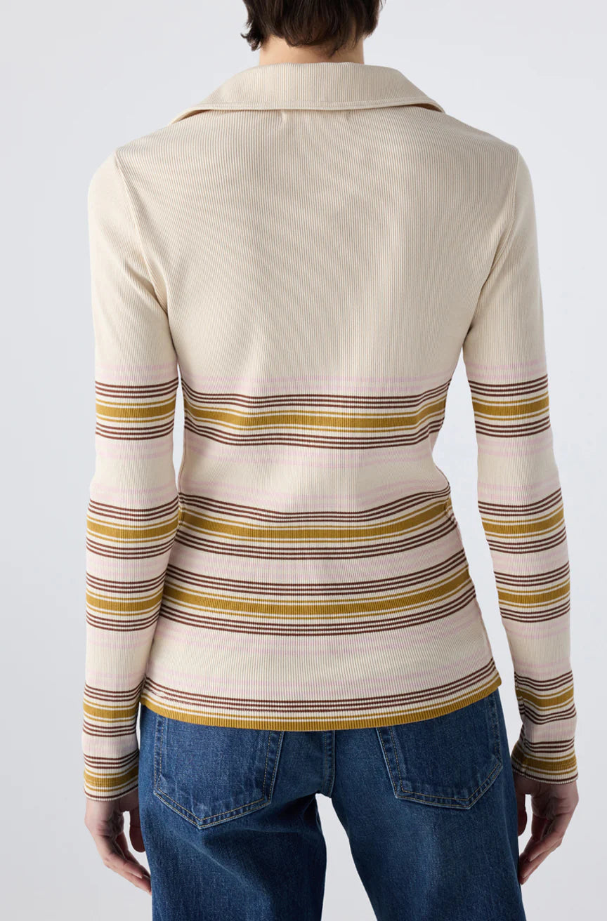 A person facing away from the camera wears the Desiree Tee by AMO Denim, a cream-colored ribbed top with horizontal pink, yellow, and brown stripes and a classic collar, paired with blue jeans against a plain background.
