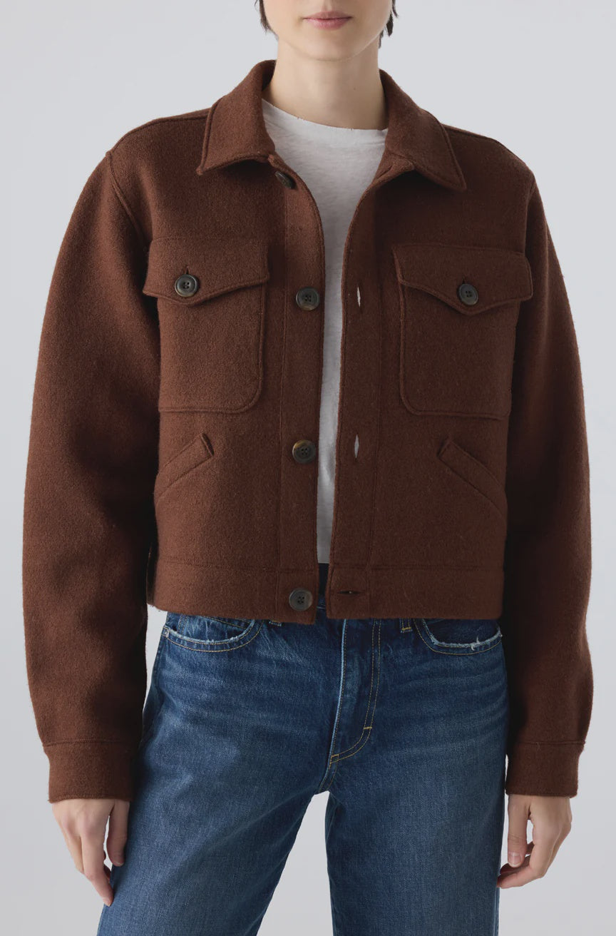 A model showcases the AMO Denim Isabelle Jacket, a cropped brown wool trucker with front pockets, styled over a white shirt and blue jeans, standing against a plain light background for a flattering silhouette.