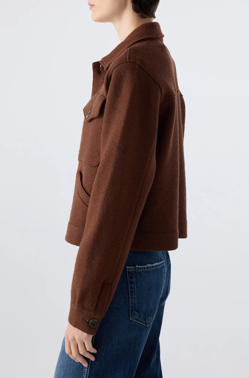 Model wearing the AMO Denim Isabelle Jacket in brown wool with flap pockets and buttoned cuffs, paired with blue jeans, shown from the side against a plain light background for a flattering silhouette.