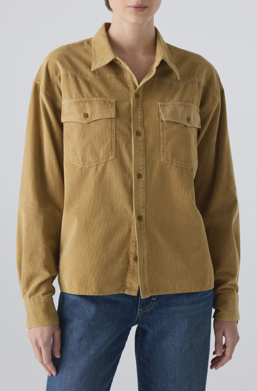 A person wearing the AMO Denim Jenn Shirt—a loose-fitting, tan corduroy top with front pockets and long sleeves—paired with blue jeans, stands against a plain light background.