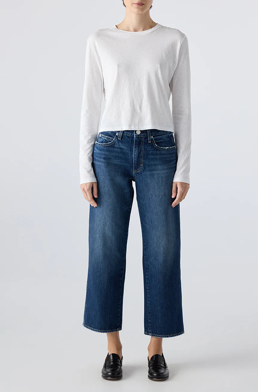 A person stands facing forward wearing a plain white long-sleeve shirt, AMO Denim Billie Pants—premium rigid denim cropped wide leg jeans—and black slip-on shoes against a light grey background.