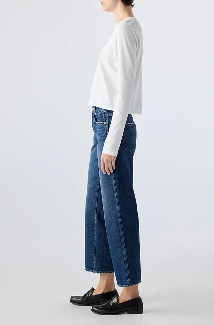 A person stands in profile wearing a long-sleeve white top, AMO Denim Billie Pants in premium rigid denim with a cropped wide leg, and black loafers against a plain light background.