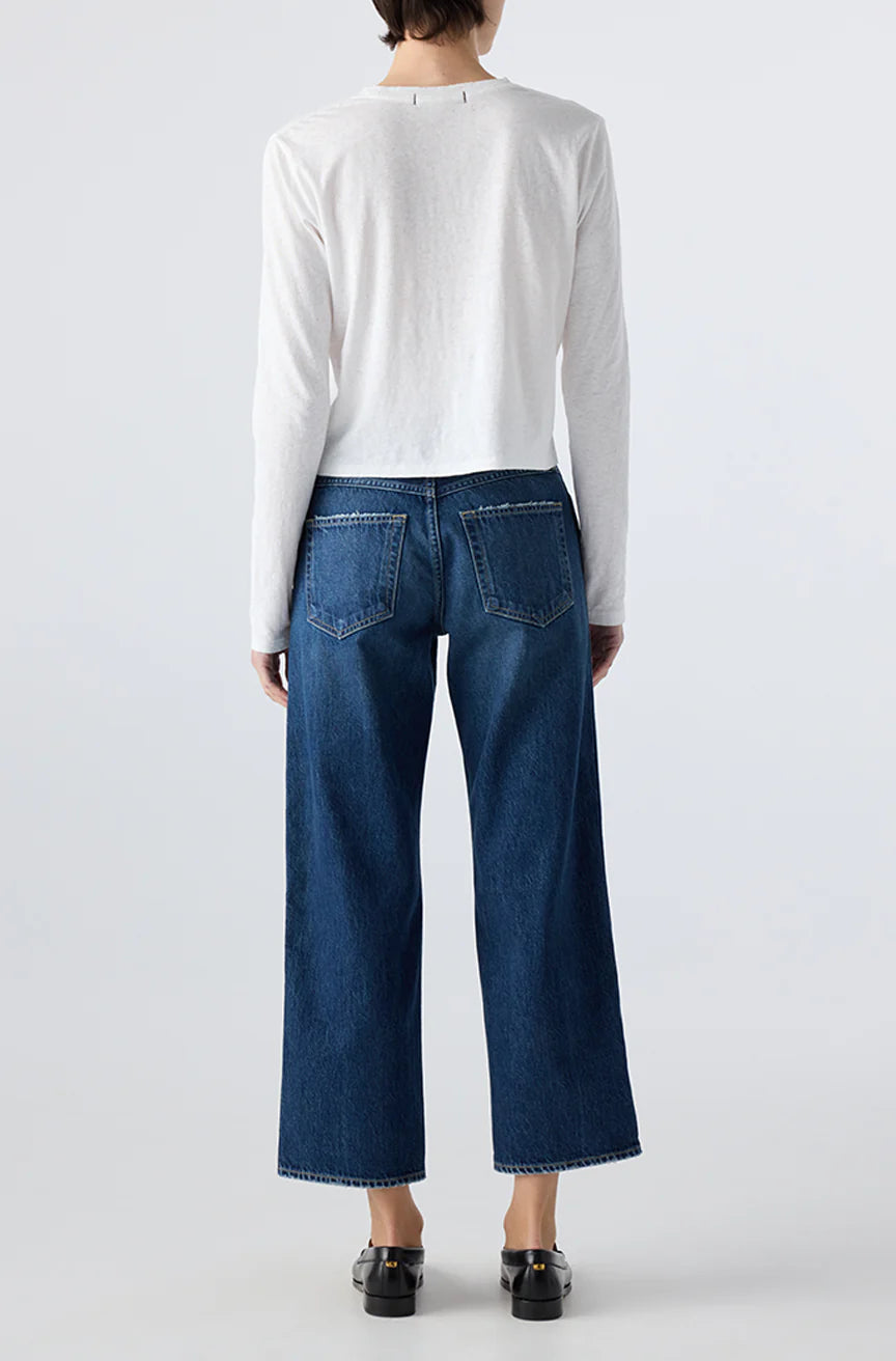 A person faces away from the camera, wearing a long-sleeved white top and AMO Denim Billie Pants—premium rigid denim cropped wide-leg jeans—with black shoes, set against a plain light background.