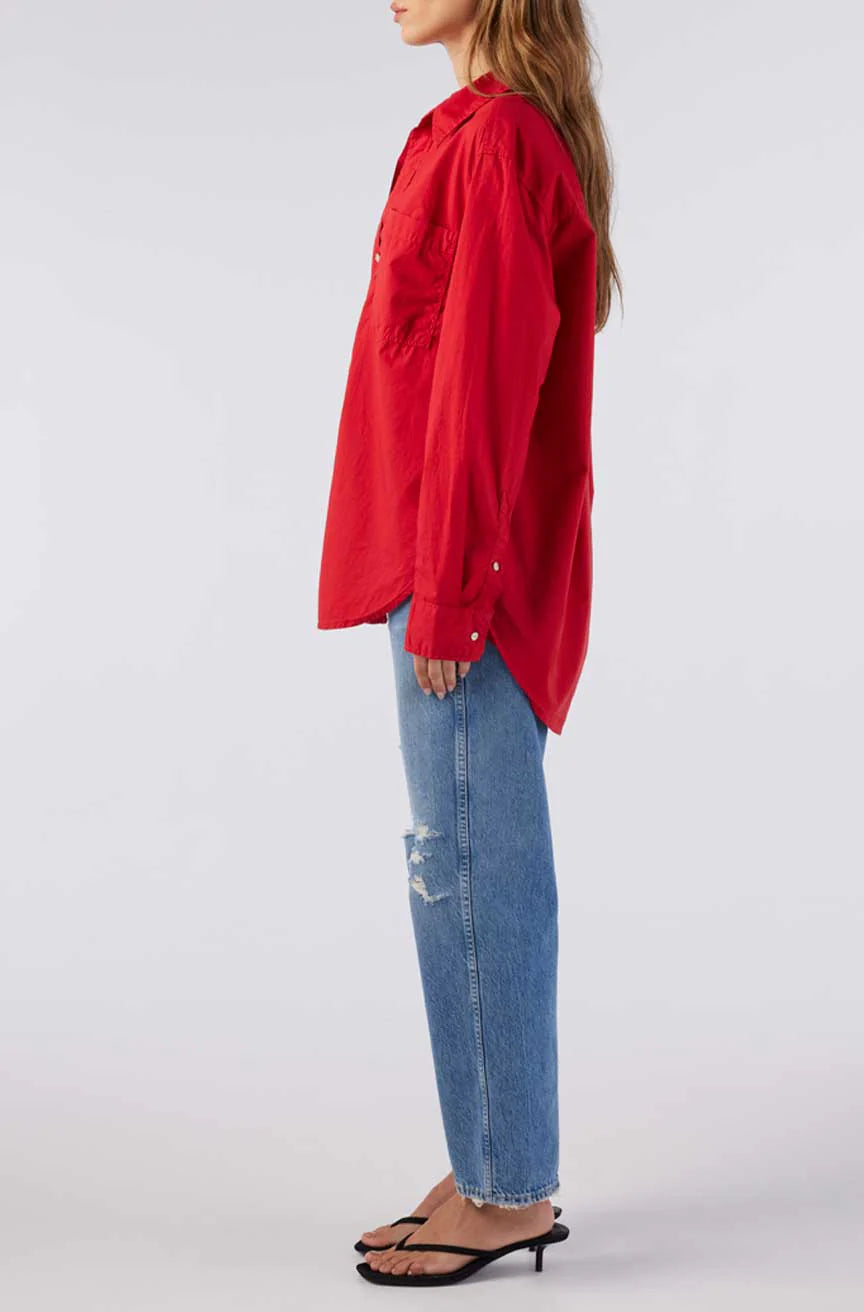 A woman in a Ruth Oversized Shirt by AMO Denim, featuring a red vintage wash, and blue ripped jeans stands in profile. She pairs the look with black heeled sandals against a plain white background.