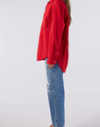 A woman in a Ruth Oversized Shirt by AMO Denim, featuring a red vintage wash, and blue ripped jeans stands in profile. She pairs the look with black heeled sandals against a plain white background.