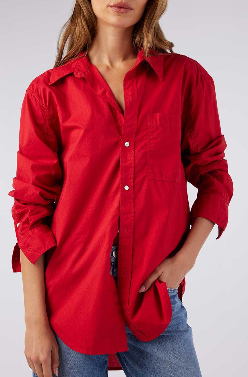 A person wearing AMO Denim's Ruth Oversized Shirt, bright red and vintage-inspired with sleeves rolled up, styled open-necked. Their hand is tucked into the pocket of stonewashed cotton poplin jeans. The face remains unseen.