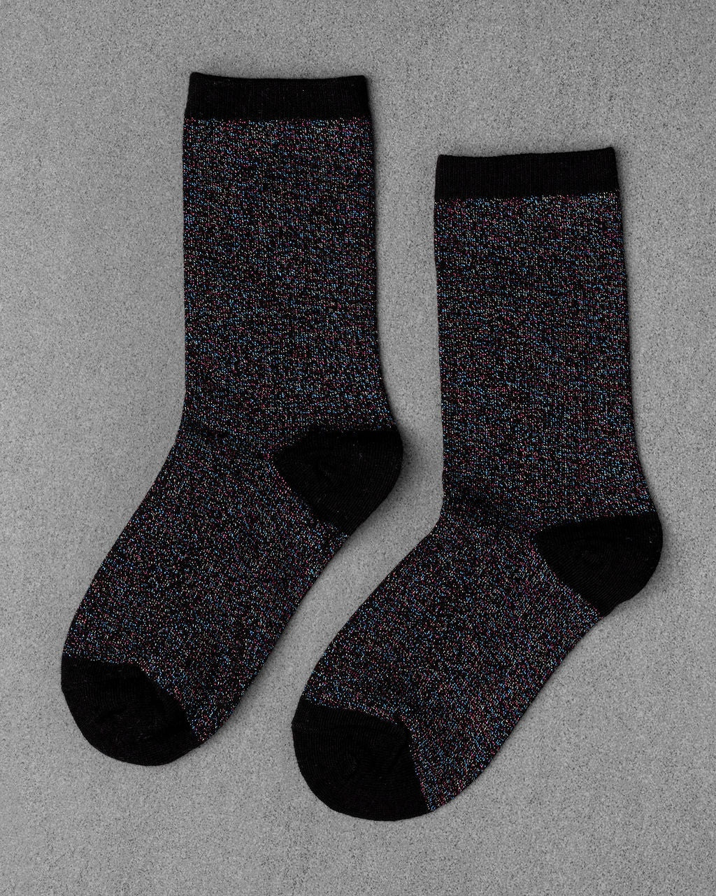 A pair of Unified Glitter Socks featuring a black base with multicolored speckles, plus black toes, heels, and cuffs, is laid flat on a gray surface.