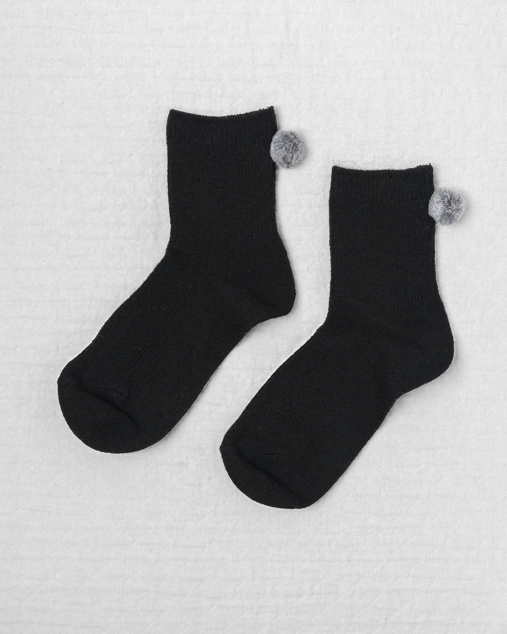 Unified's Winter Pom Pom Socks are cozy black ankle socks featuring small gray pompoms near the top cuffs, shown on a light textured surface.