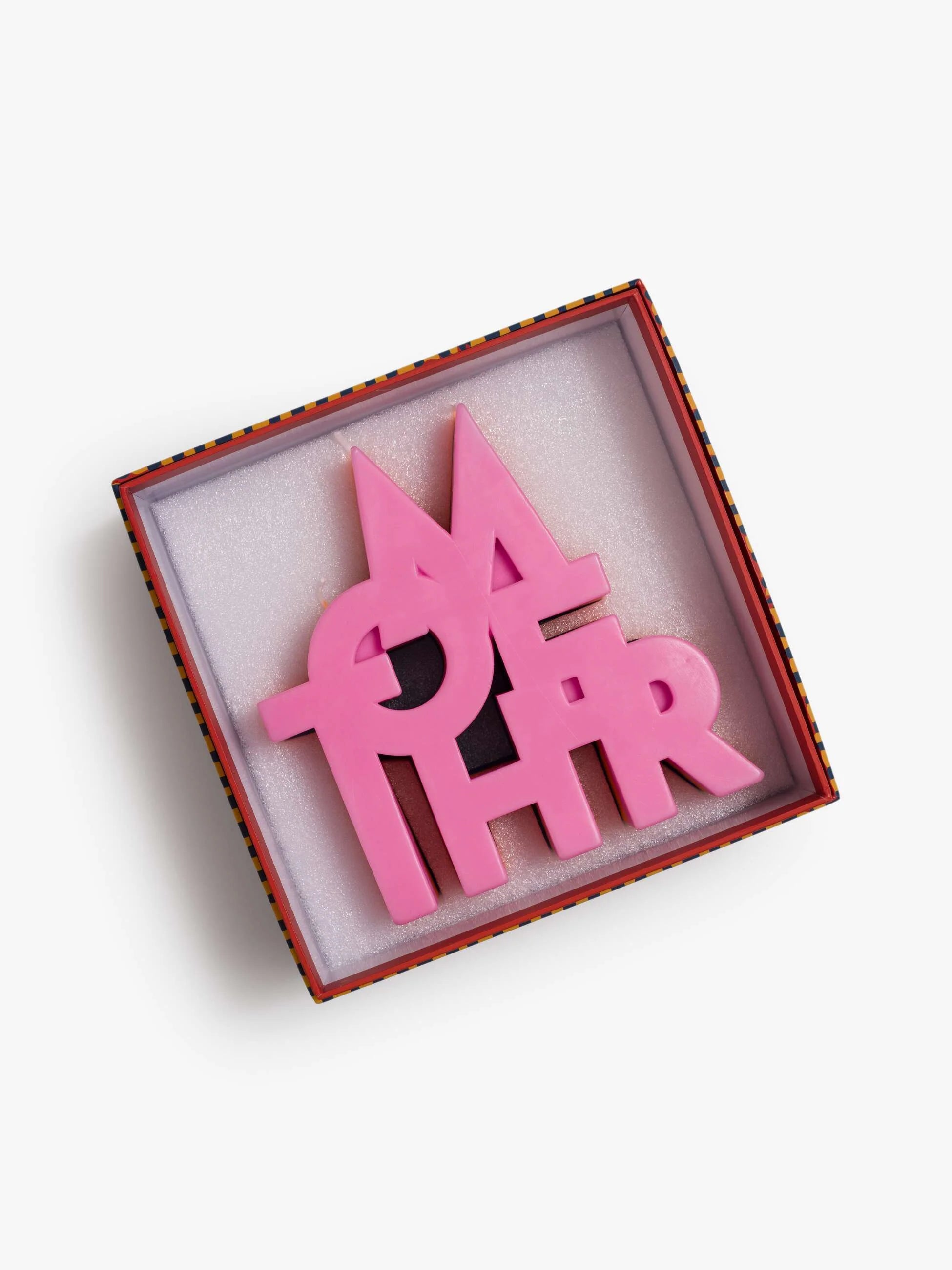 The Mother Burnout Candle by Mother features bold, overlapping letters spelling "MOTHER" in pink soy wax and comes in a square gift box lined with white padding. Available in Blue, Orange, and Pink.