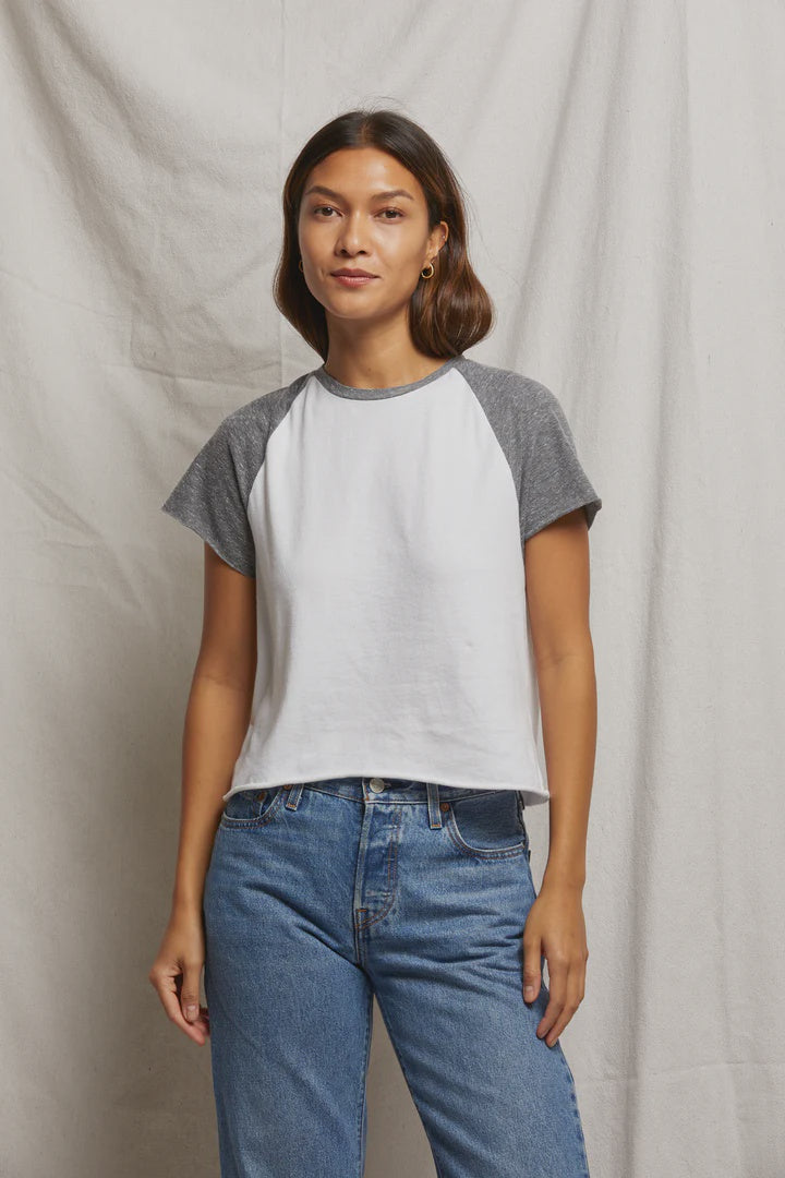 A woman poses before a white backdrop in a Perfectwhitetee Britt SS Raglan Tee with gray sleeves and blue jeans. Her long brown hair and subtle earrings complement her gentle smile and hands by her sides, radiating retro charm.