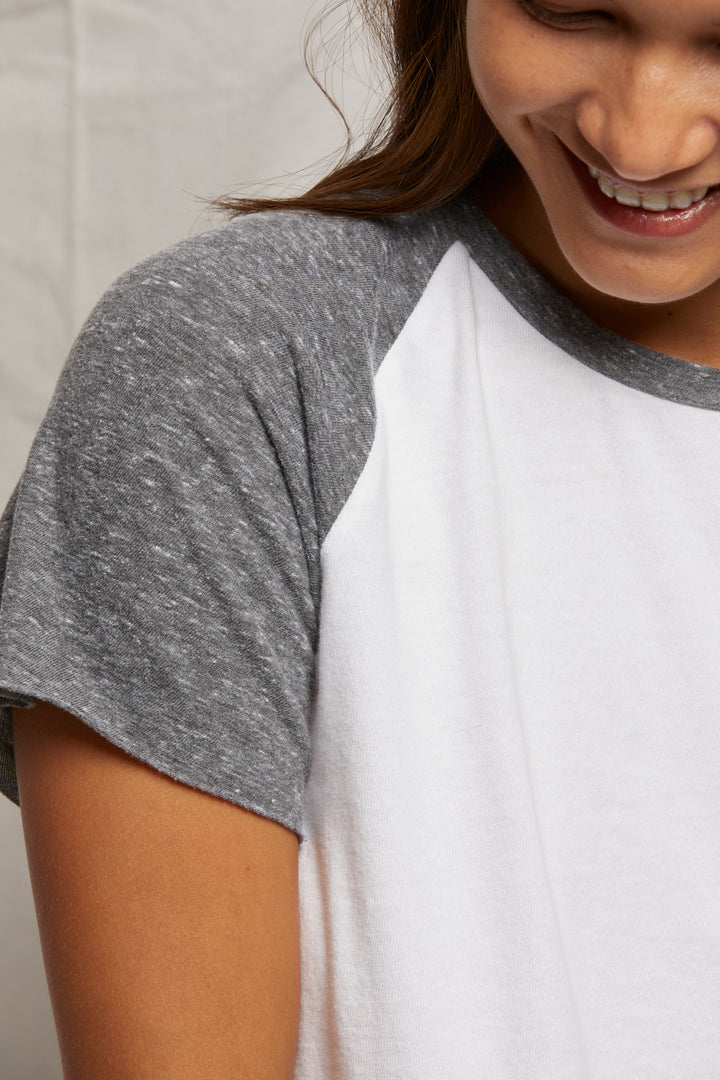 A person smiles in a Perfectwhitetee Britt SS Raglan Tee, a short-sleeved gray and white shirt with a timeless look, against a plain light fabric backdrop.