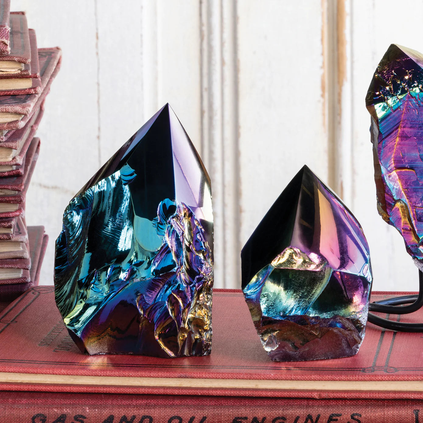 The Aura Black Obsidian Points by Blue Ocean Traders feature iridescent crystal points with metallic blue, purple, and gold hues, displayed on a red book among vintage books—a captivating sculptural accent for any space.