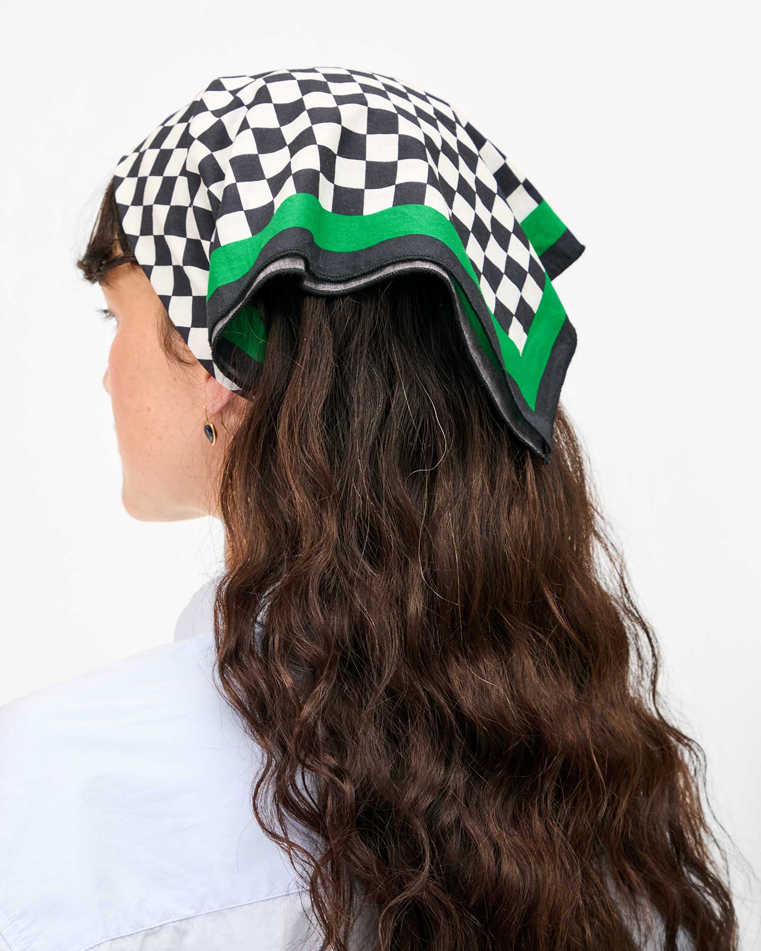 A person with long, wavy brown hair wears a Clare V Bandana by Clare Vivier, a cotton black and white checkered scarf with green edges, stylishly tied over their head. They sport a light blue shirt and face away against a plain white background.