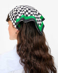 A person with long, wavy brown hair wears a Clare V Bandana by Clare Vivier, a cotton black and white checkered scarf with green edges, stylishly tied over their head. They sport a light blue shirt and face away against a plain white background.