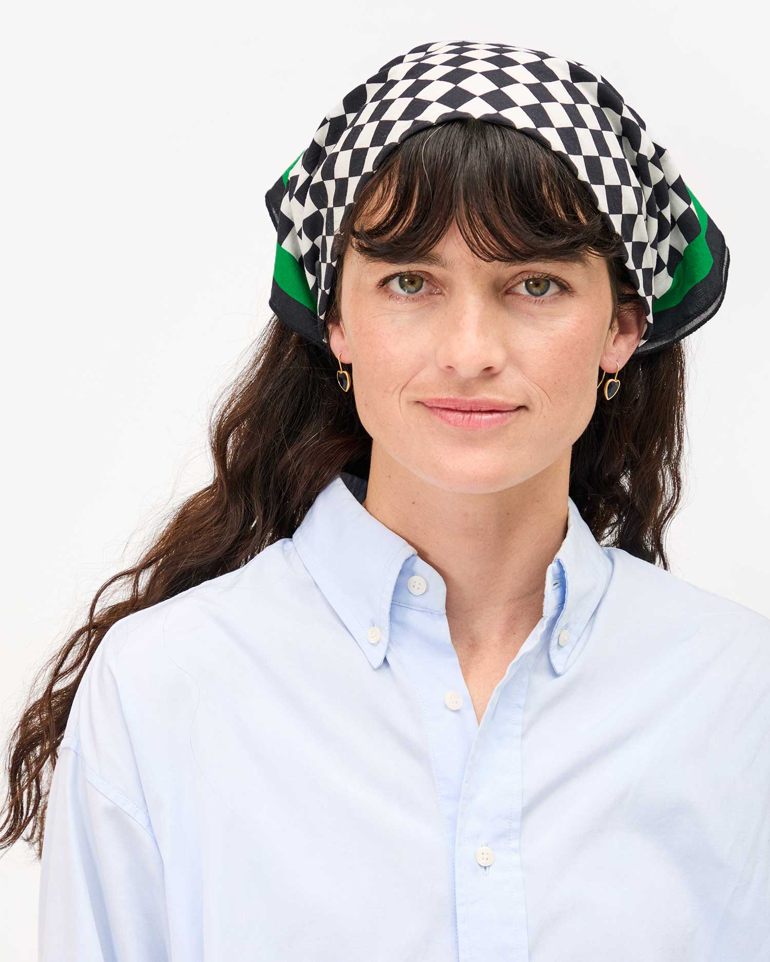 A woman with long dark hair wearing a blue button-up shirt smiles softly at the camera, showcasing a Clare V Bandana by Clare Vivier, a black and white checkered cotton headscarf with green accents, made in India, all set against a plain white background.