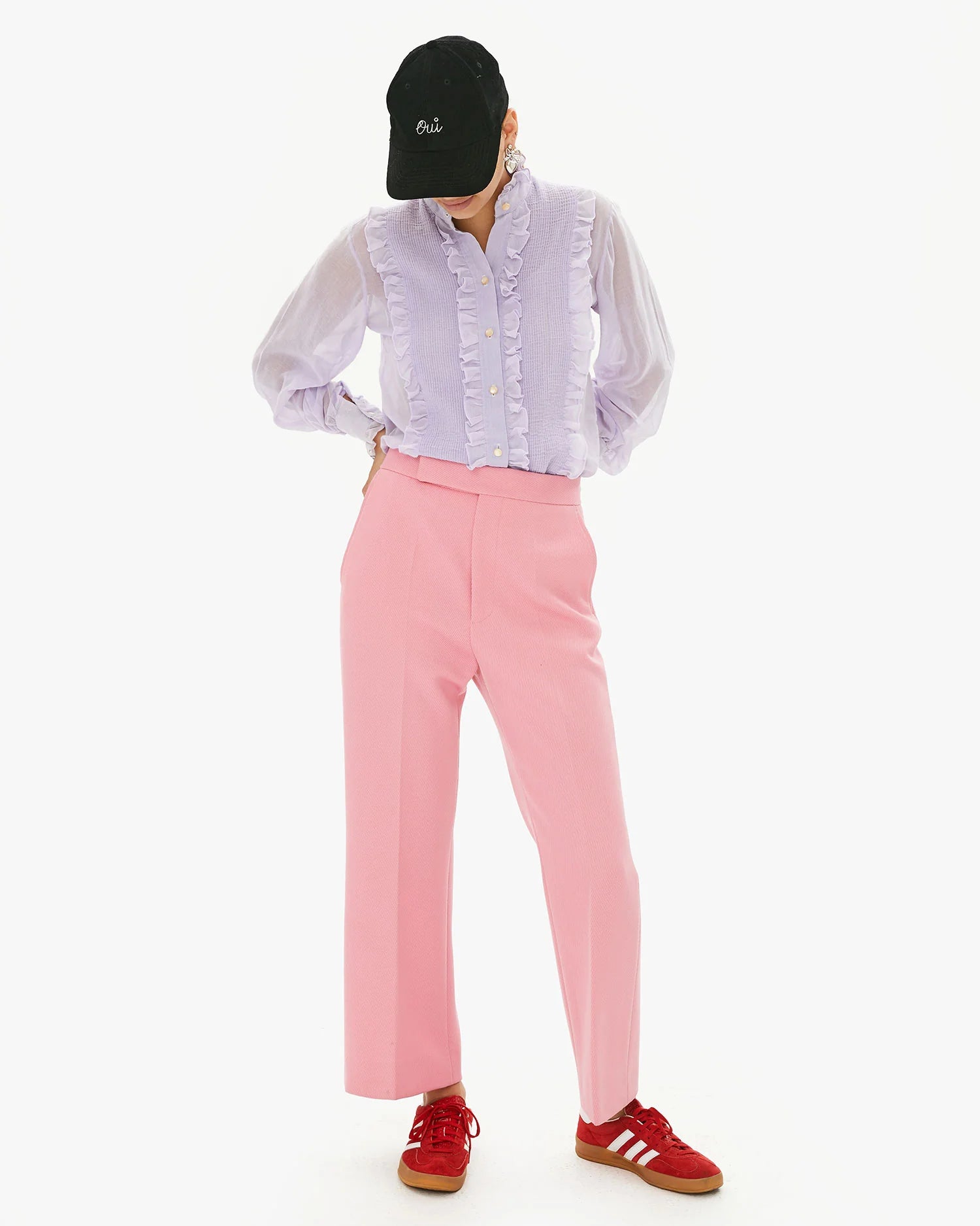 A person wearing a black Clare Vivier Baseball Hat resembles a celebrity in disguise, sporting a lilac ruffled blouse, pink wide-leg pants, and red sneakers. They stand against a plain white background with hands in pockets and head slightly tilted down.