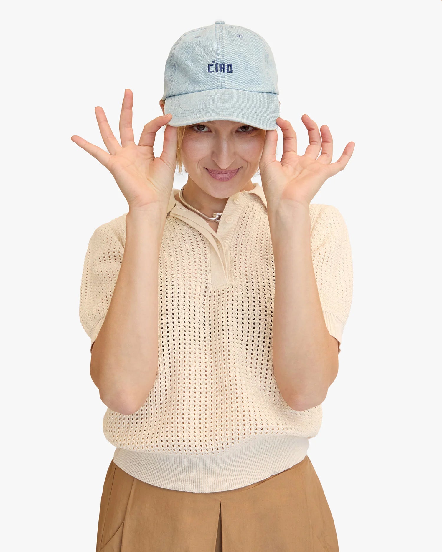 Wearing Clare Vivier's embroidered denim Baseball Hat with "Ciao," a person playfully adjusts the brim. They sport a knit beige short-sleeve shirt and tan pants while subtly smiling against a plain white background, enhancing their celebrity-in-disguise charm.