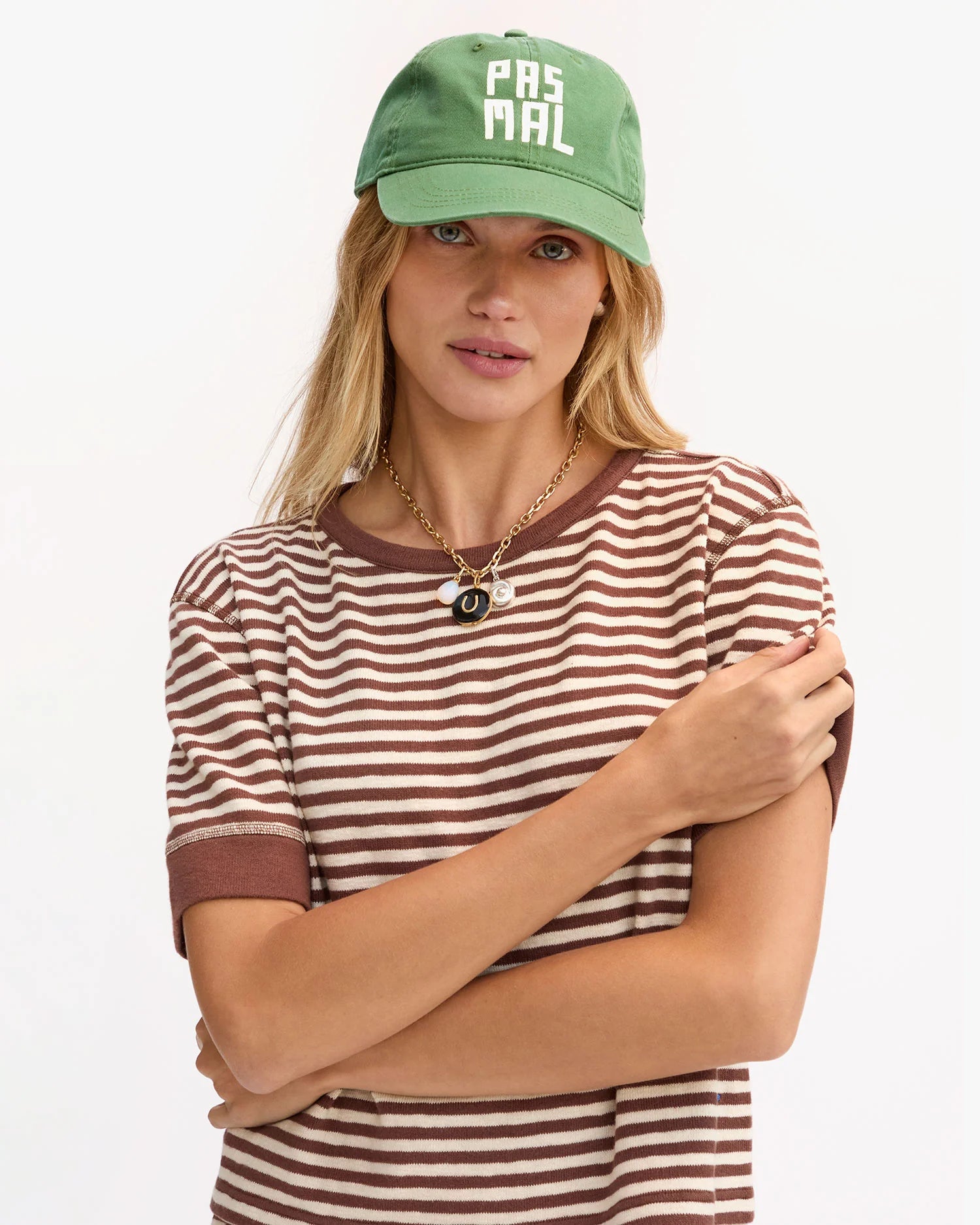 A woman wears the Clare Vivier Baseball Hat PasMal, featuring "PAS MAL" text and an adjustable metal closure, paired with a 100% cotton brown-and-white striped tee and layered necklaces, standing with arms crossed against a white background.