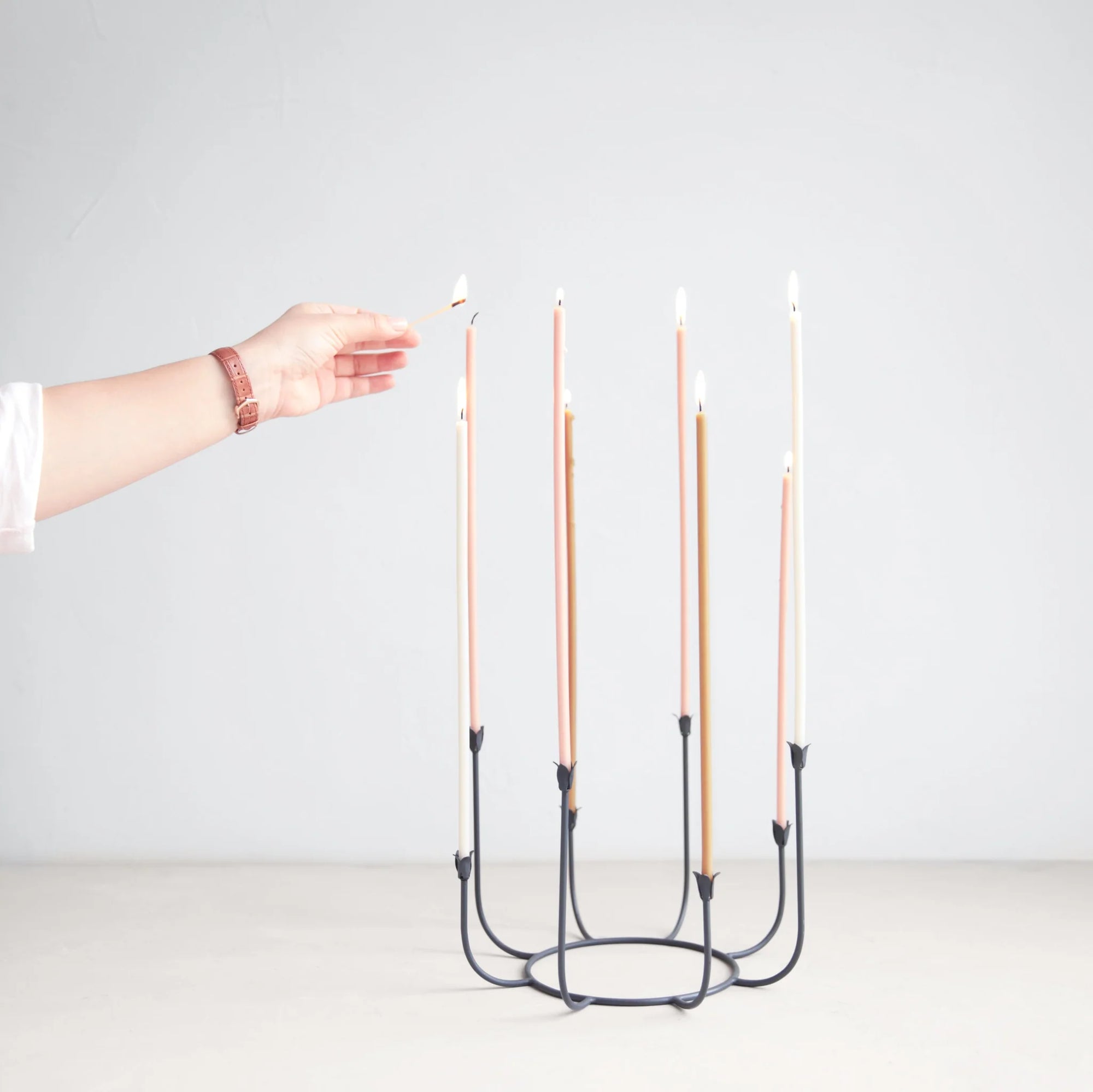 A hand is lighting the final candle on a Beacon Long Stem Candelabra by The Floral Society. These handmade tapers, presented in various shades of pink and beige, are elegantly displayed against a simple, light gray background.