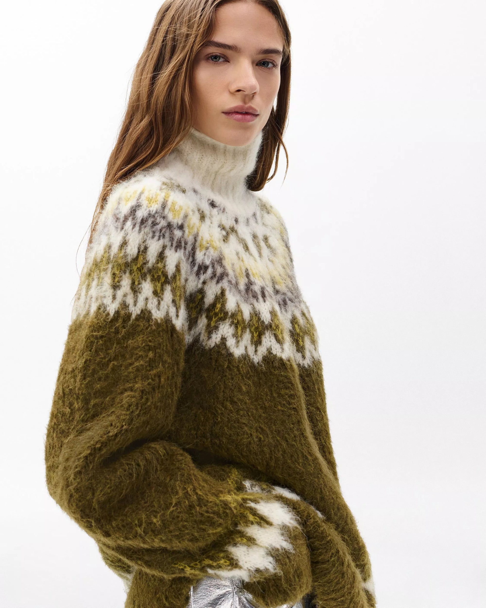 A woman with long brown hair models the Rag & Bone beth oversized fair isle tneck sweater in olive green, white, and gray, standing against a plain white background.