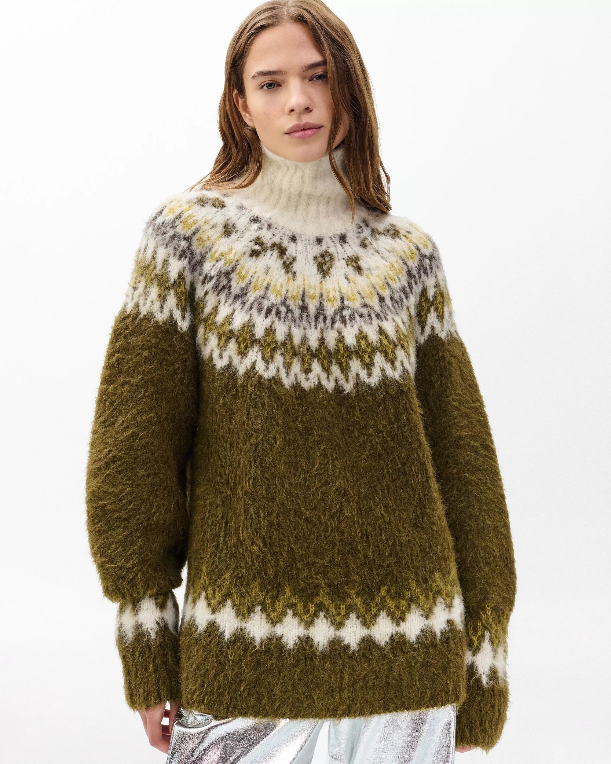 A woman with long brown hair models the Beth Oversized Fair Isle Tneck by Rag & Bone, featuring an olive green and white pattern and high neck, standing against a plain white background.