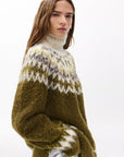 A woman with long brown hair models the Rag & Bone beth oversized fair isle tneck sweater in olive green, white, and gray, standing against a plain white background.