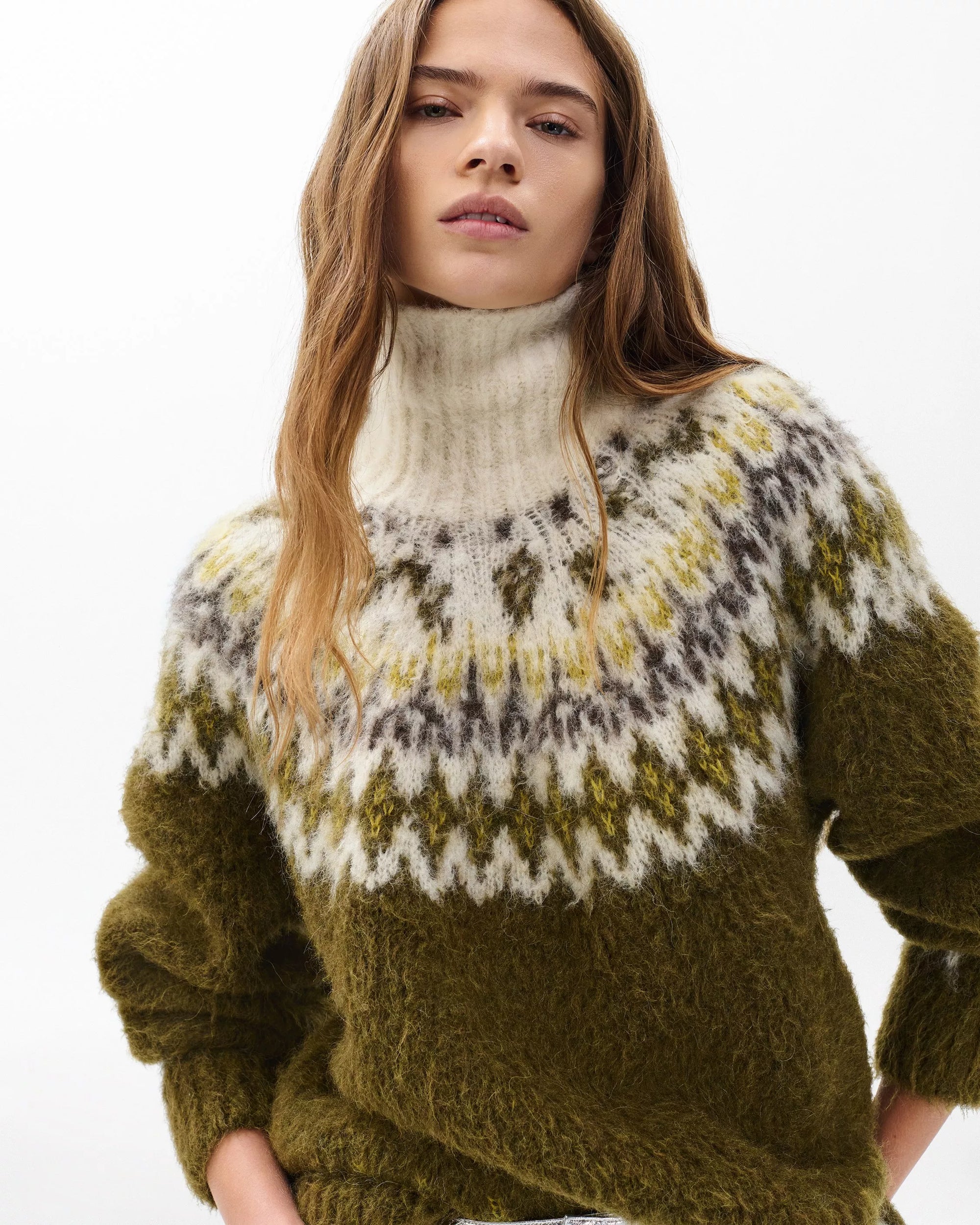 A woman with long, wavy brown hair wears the Rag & Bone beth oversized fair isle tneck sweater with a white, gray, and yellow patterned yoke, standing confidently against a plain white background.