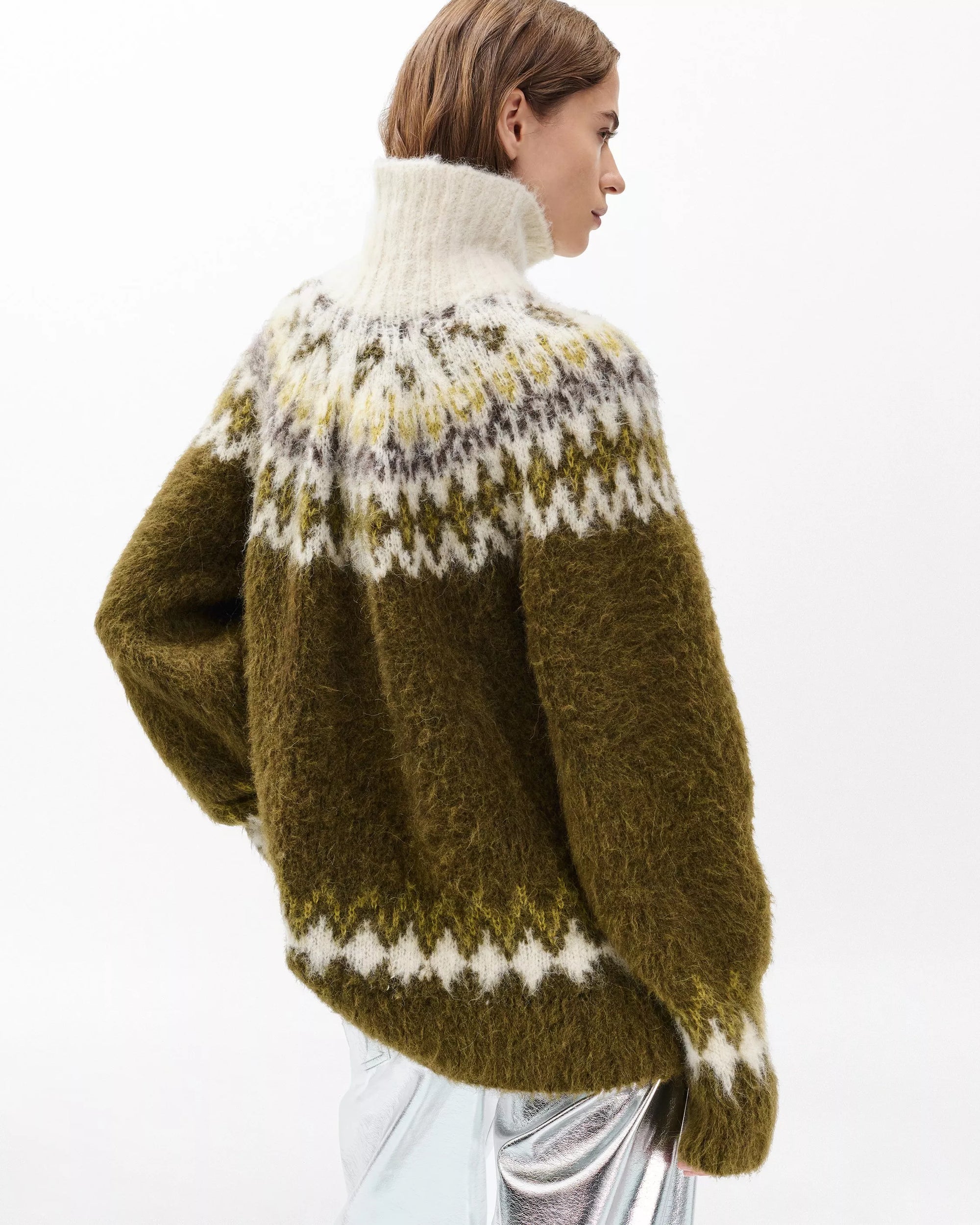 A person with short brown hair wears the Rag & Bone beth oversized fair isle tneck, a chunky knit wool sweater in olive, white, yellow, and gray with a high collar, paired with a metallic silver skirt and turned slightly to the side.