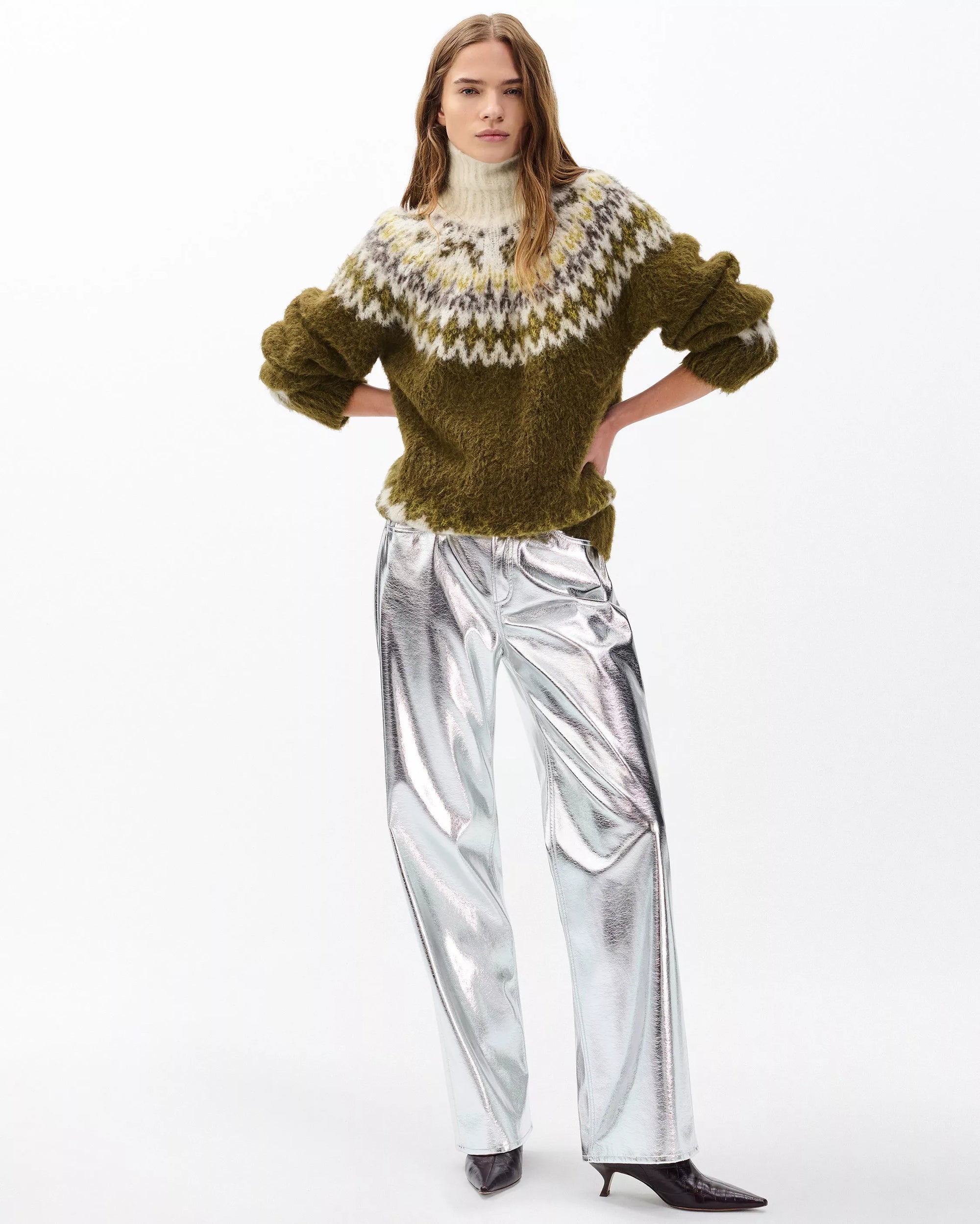 A woman stands against a white background wearing the Rag & Bone beth oversized fair isle tneck, styled with shiny metallic silver wide-leg pants and pointed dark ankle boots. Her hands are on her hips and her hair is loose.