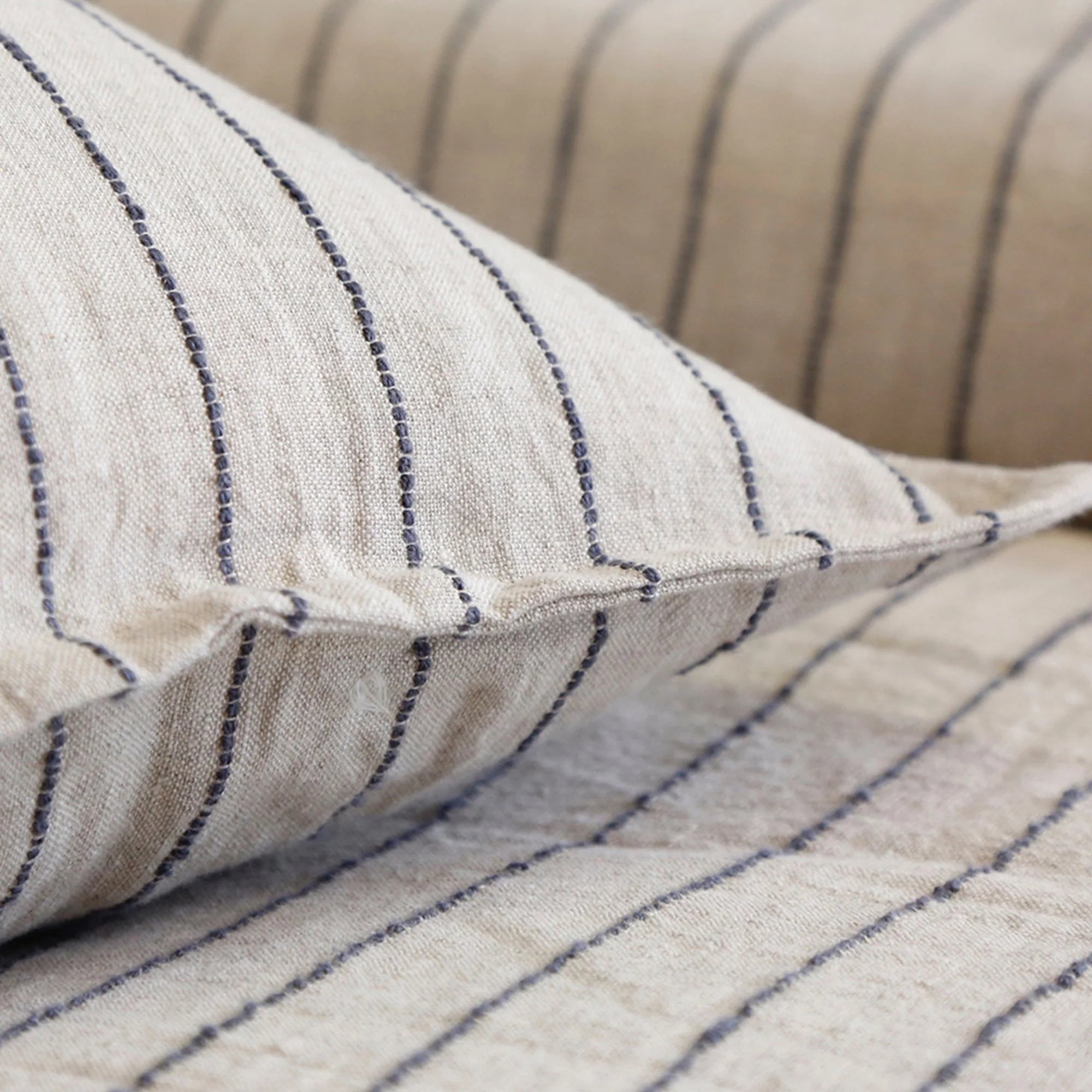 Close-up of beige Blake Big Pillow 28x36 by POM POM, featuring black vertical stripes that accentuate the hand-loomed texture and woven pattern. Part of the same style, another similarly textured surface is interwoven in the background.