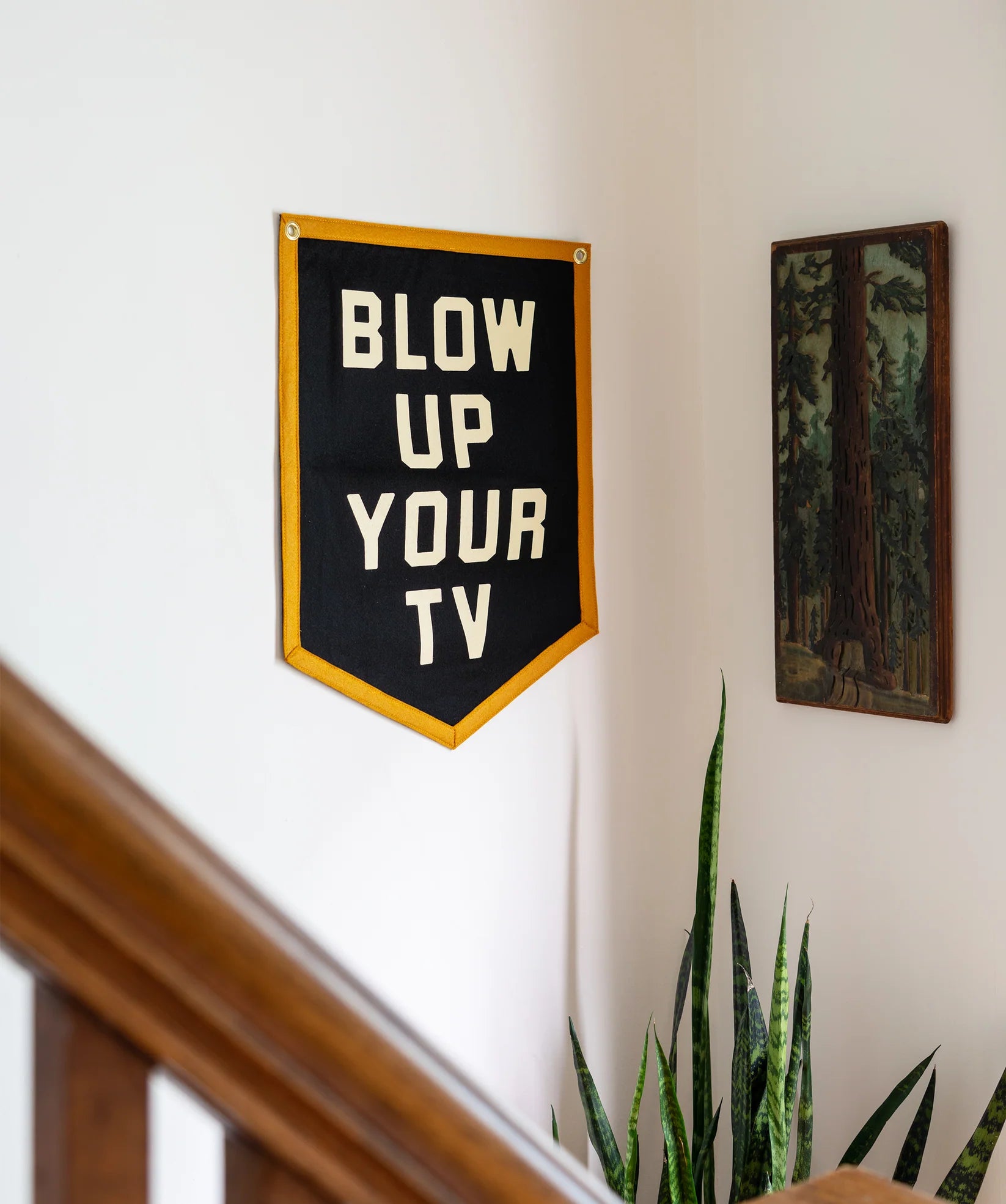 The Blow Up Your TV Pennant 18x24 by Oxford Pennant, a black flag with yellow trim and bold white text, hangs on a white wall near a painting of trees, above a potted plant and wooden staircase railing.