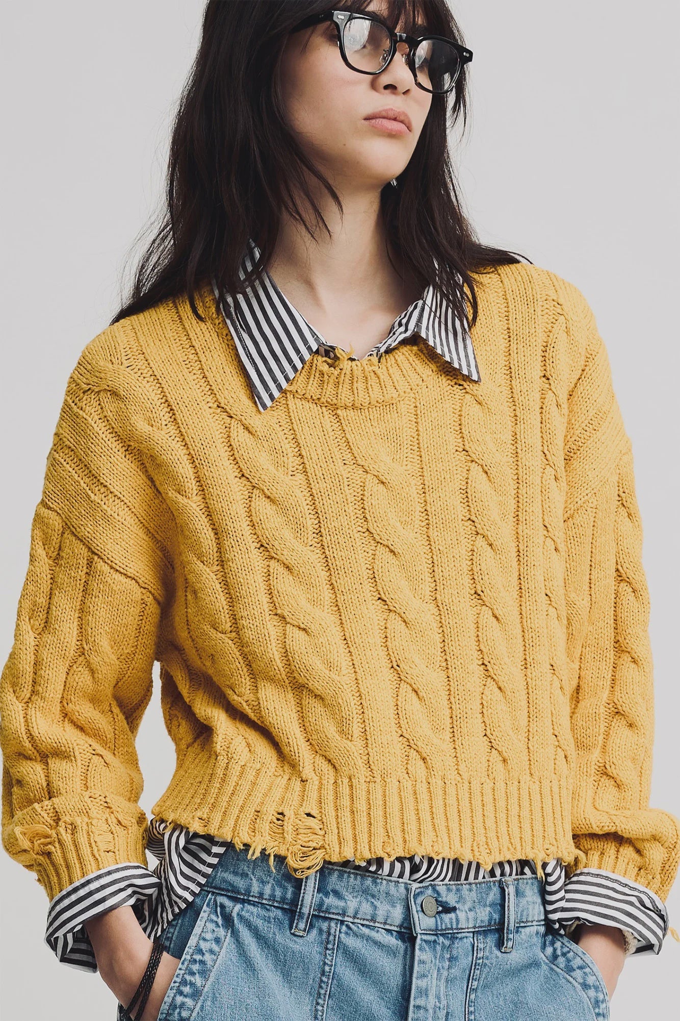 A person in the Denimist Boxy Cable Sweater, a chunky yellow cotton knit, layered over a black and white striped shirt with light blue high-waisted jeans. They have long dark hair and wear black glasses.