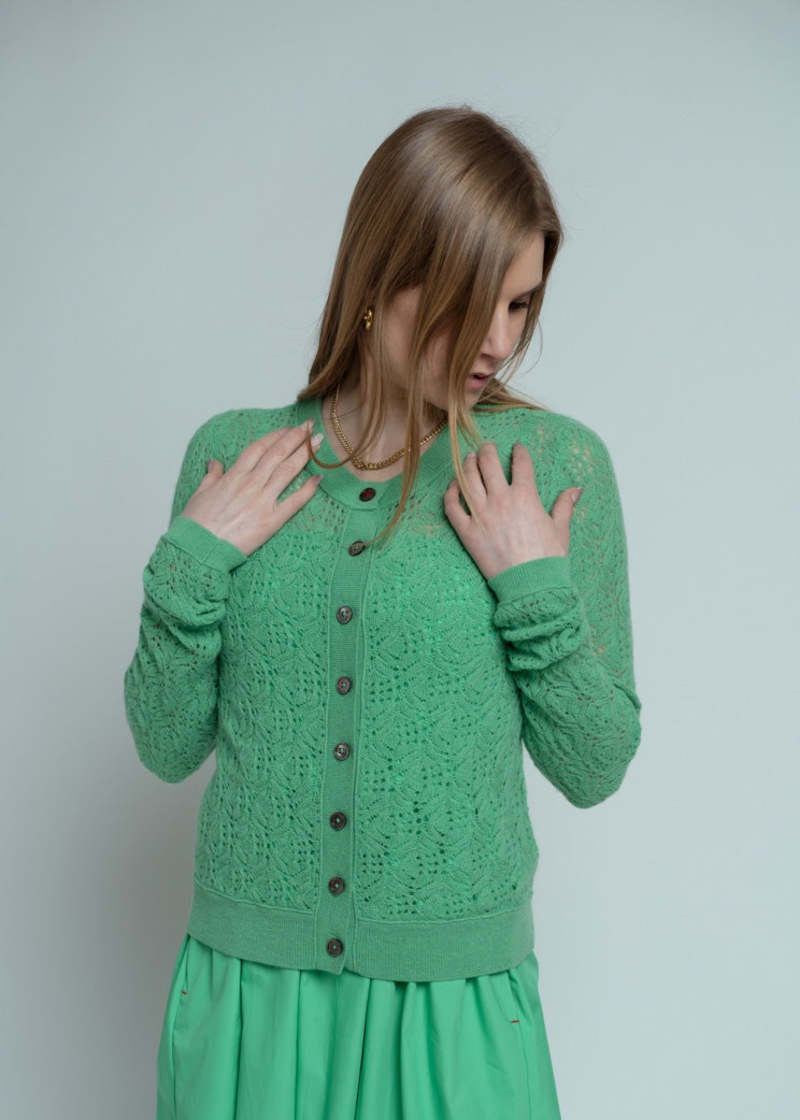 A woman with long light brown hair wears the Brazeau Tricot Chunky Perforated Cardigan over a matching dress, standing against a plain light background and touching her collar with both hands.