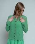 A woman with long light brown hair wears the Brazeau Tricot Chunky Perforated Cardigan over a matching dress, standing against a plain light background and touching her collar with both hands.