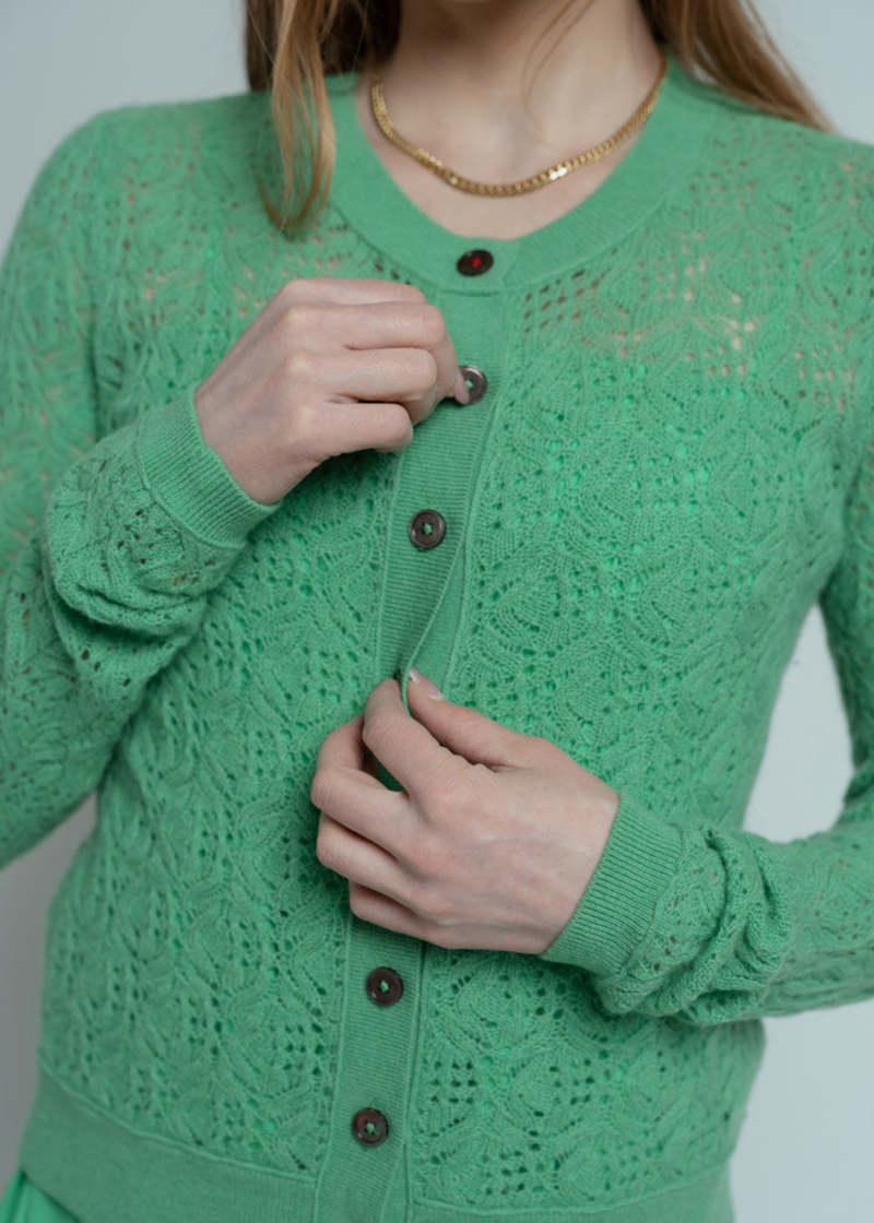 A person wearing a Brazeau Tricot Chunky Perforated Cardigan in green and a gold chain necklace is fastening a button. The image is cropped to show only the torso and hands.