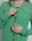 A person wearing a Brazeau Tricot Chunky Perforated Cardigan in green and a gold chain necklace is fastening a button. The image is cropped to show only the torso and hands.