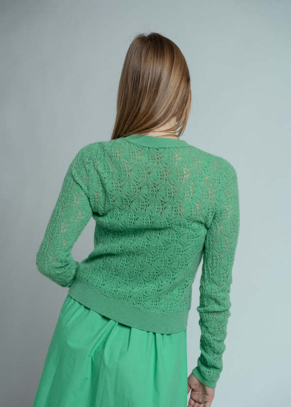 A woman with long, straight, light brown hair stands facing away, wearing the Brazeau Tricot Chunky Perforated Cardigan in green with a matching skirt against a plain light gray background.