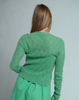 A woman with long, straight, light brown hair stands facing away, wearing the Brazeau Tricot Chunky Perforated Cardigan in green with a matching skirt against a plain light gray background.