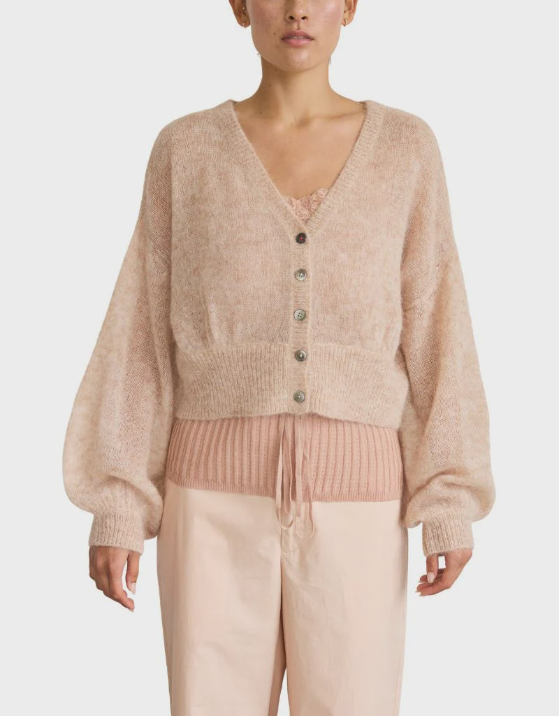 A woman wears a soft, light beige Whisper Cardigan by Brazeau Tricot over a ribbed blush pink knit top and light beige high-waisted pants, standing against a plain white background.