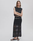 A woman with wavy brown hair wears the Rag & Bone britt maxi skirt—a sheer black crochet summer skirt with a belt—paired with a sleeveless black top and black sandals, posing confidently against a plain white background.
