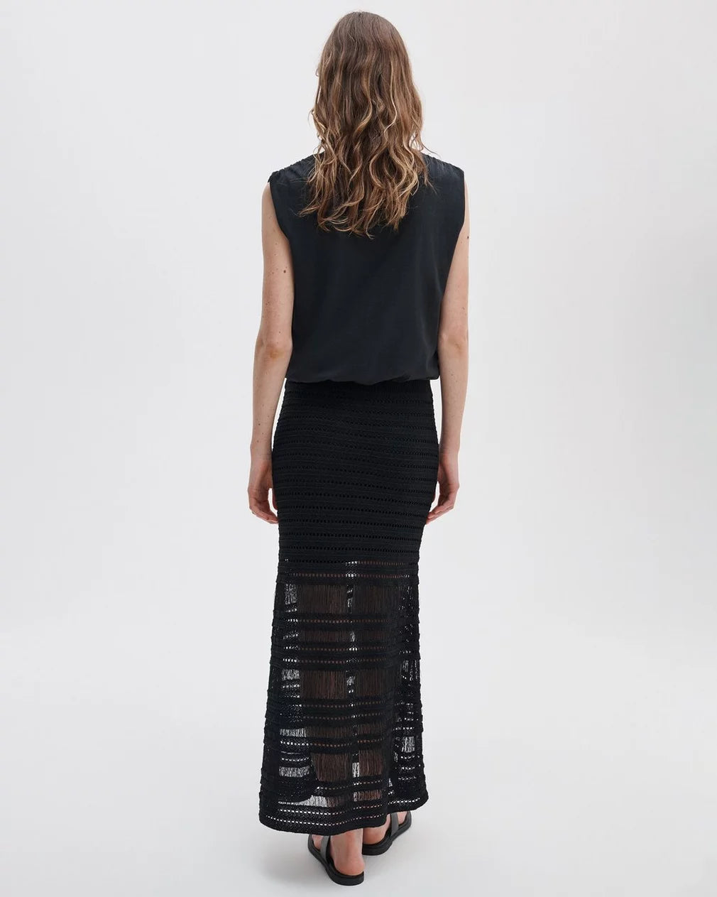 A woman with wavy brown hair stands facing away in a sleeveless black top and the Rag & Bone britt maxi skirt, a summer-ready, semi-sheer black crochet piece with horizontal patterns. The background is plain and light-colored.