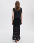 A woman with wavy brown hair stands facing away in a sleeveless black top and the Rag & Bone britt maxi skirt, a summer-ready, semi-sheer black crochet piece with horizontal patterns. The background is plain and light-colored.