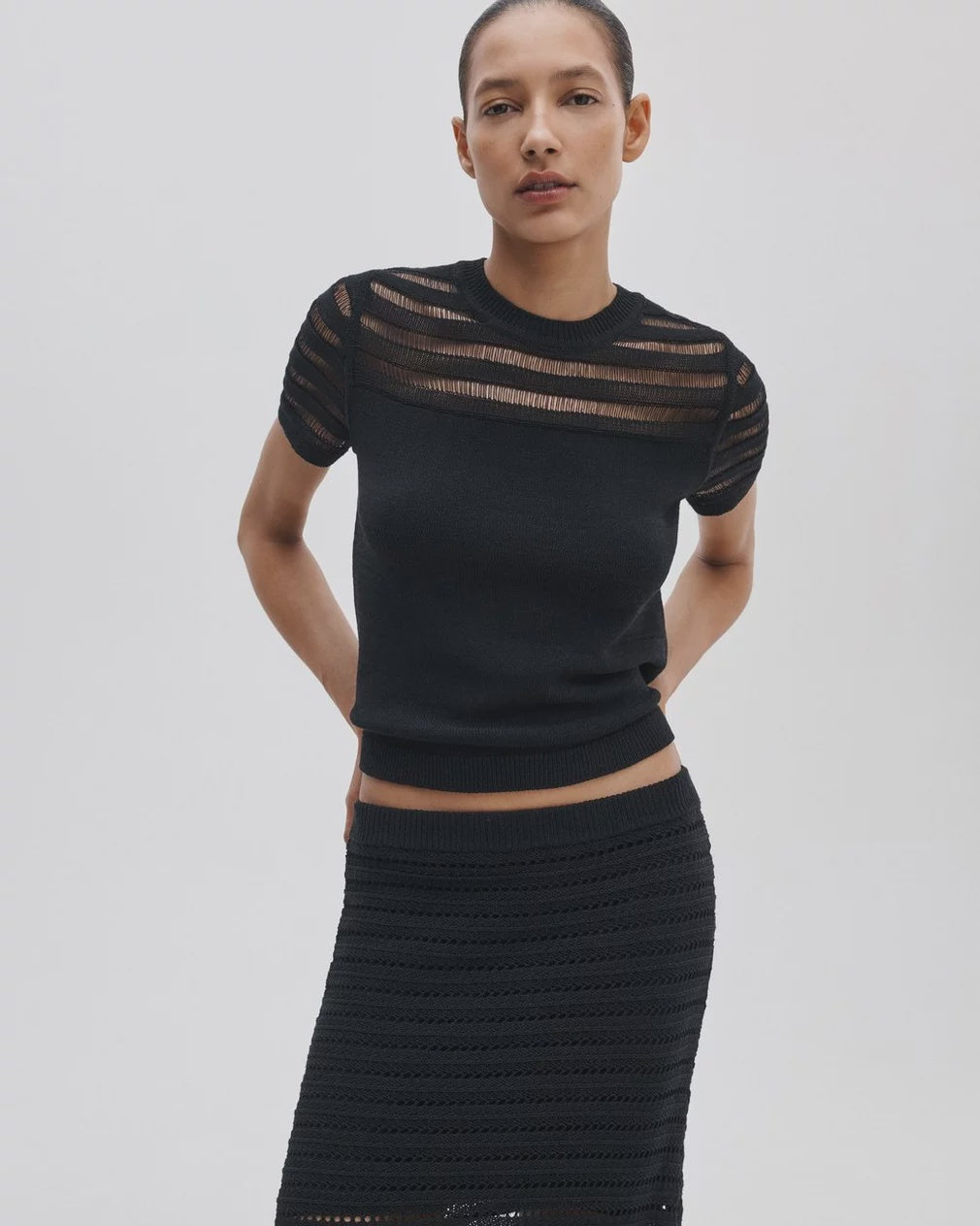 A woman poses against a light background in the Rag & Bone britt ss crew, an airy striped summer sweater with sheer details at the shoulders and sleeves, paired with a matching black textured skirt.