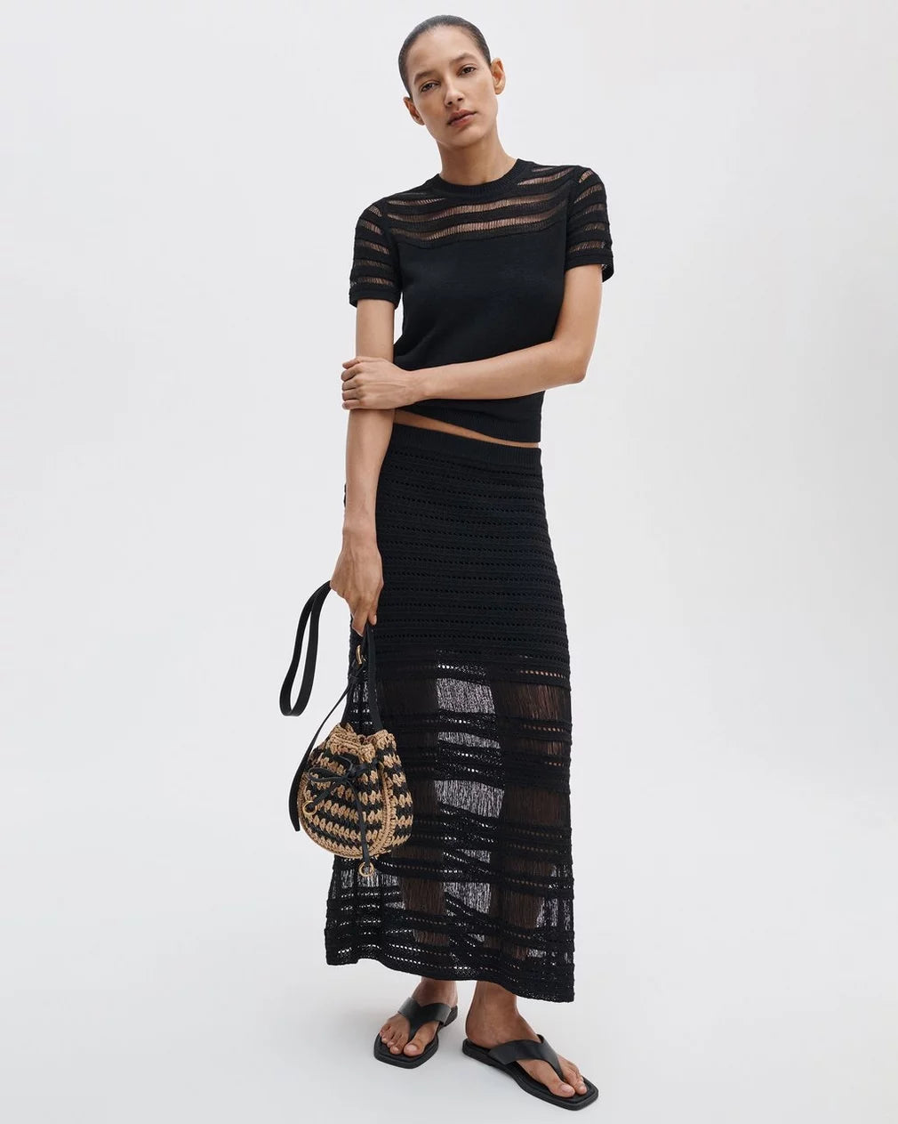 A woman wears the Rag & Bone britt ss crew, a black sheer short-sleeve top, paired with a matching long crochet skirt, black sandals, woven bag, and lightweight cotton sweater against a plain background.