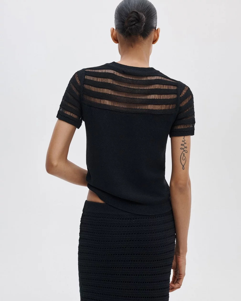 A person with their hair in a bun is seen from behind, wearing the Rag & Bone britt ss crew— a short-sleeve striped sweater with sheer horizontal stripes on the upper back—paired with a black textured skirt. A geometric tattoo appears on their left arm.
