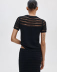 A person with their hair in a bun is seen from behind, wearing the Rag & Bone britt ss crew— a short-sleeve striped sweater with sheer horizontal stripes on the upper back—paired with a black textured skirt. A geometric tattoo appears on their left arm.