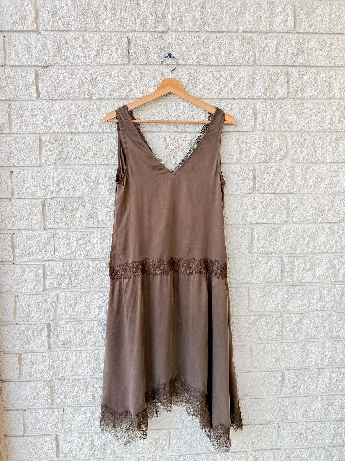 Sleeveless brown slip dress with lace trim by Xirena, cupro viscose fabric, displayed on a hanger against white brick.