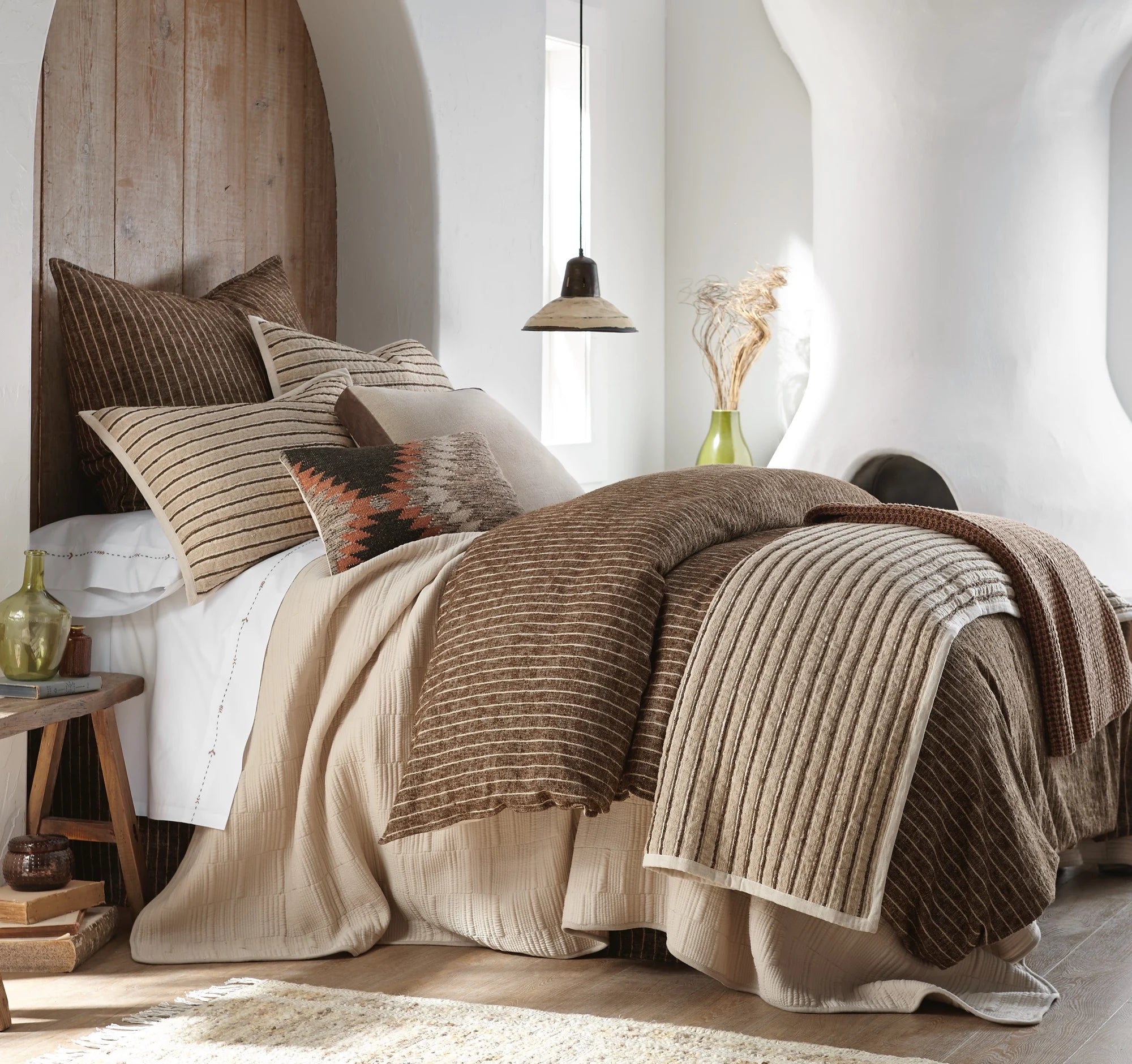 The Amity Home Bruno Duvet Cover King, featuring beige and brown stripes, styled with decorative pillows and a knit throw, brings modern elegance to a bright room with wood accents, a side table, and dried plants in a vase.