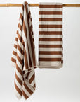 Two Maria Pool Towels by Autumn Sonata, crafted from organic cotton for luxurious absorbency, hang on a wooden rod against a plain white background. One oversized beach towel is draped loosely, while the other is neatly folded. The towels feature a broad, horizontal stripe pattern in brown and white.