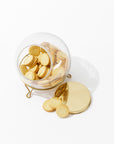 The Scullery Jar by Sir/Madam, a transparent hand-blown glass jar filled with golden cream-filled cookies, sits elegantly on a gold metal stand. The polished brass lid of the jar is off and placed beside it on the table, with some cookies laid out in front of the jar. This sophisticated kitchen storage solution is set against a white background.